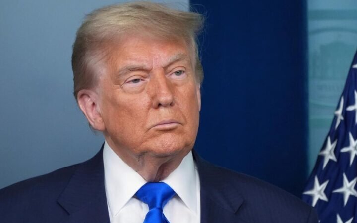 تُظهر الصورة لقطة مقربة للرئيس الأمريكي دونالد ترامب يرتدي بدلة رسمية كحلية مع قميص أبيض ورابطة عنق زرقاء وشعار علم الولايات المتحدة على ياقة بدلته، بينما يظهر جانب من العلم الأمريكي في الخلفية على جهة اليمين.