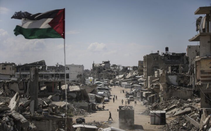 تظهر الصورة العلم الفلسطيني يرفرف فوق شارع مدمر تحيط به مبانٍ مدمرة في منطقة مشروع بيت لاهيا، شمالي قطاع غزة.